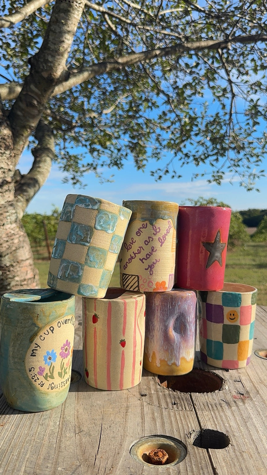 Seven colorful handmade pottery tumblers displayed outdoors beneath a leafy tree